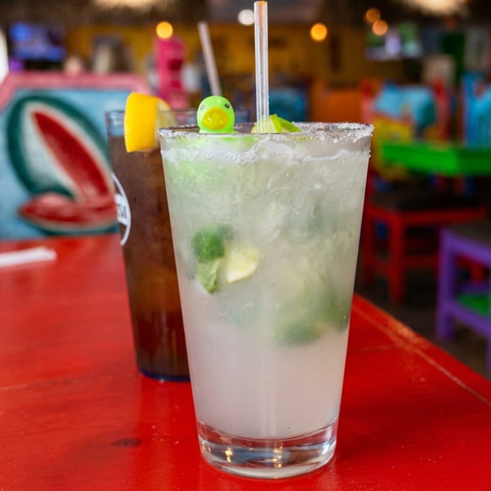 Frosty lime cocktail garnished with citrus and mint on a red table in a colorful, lively restaurant.
