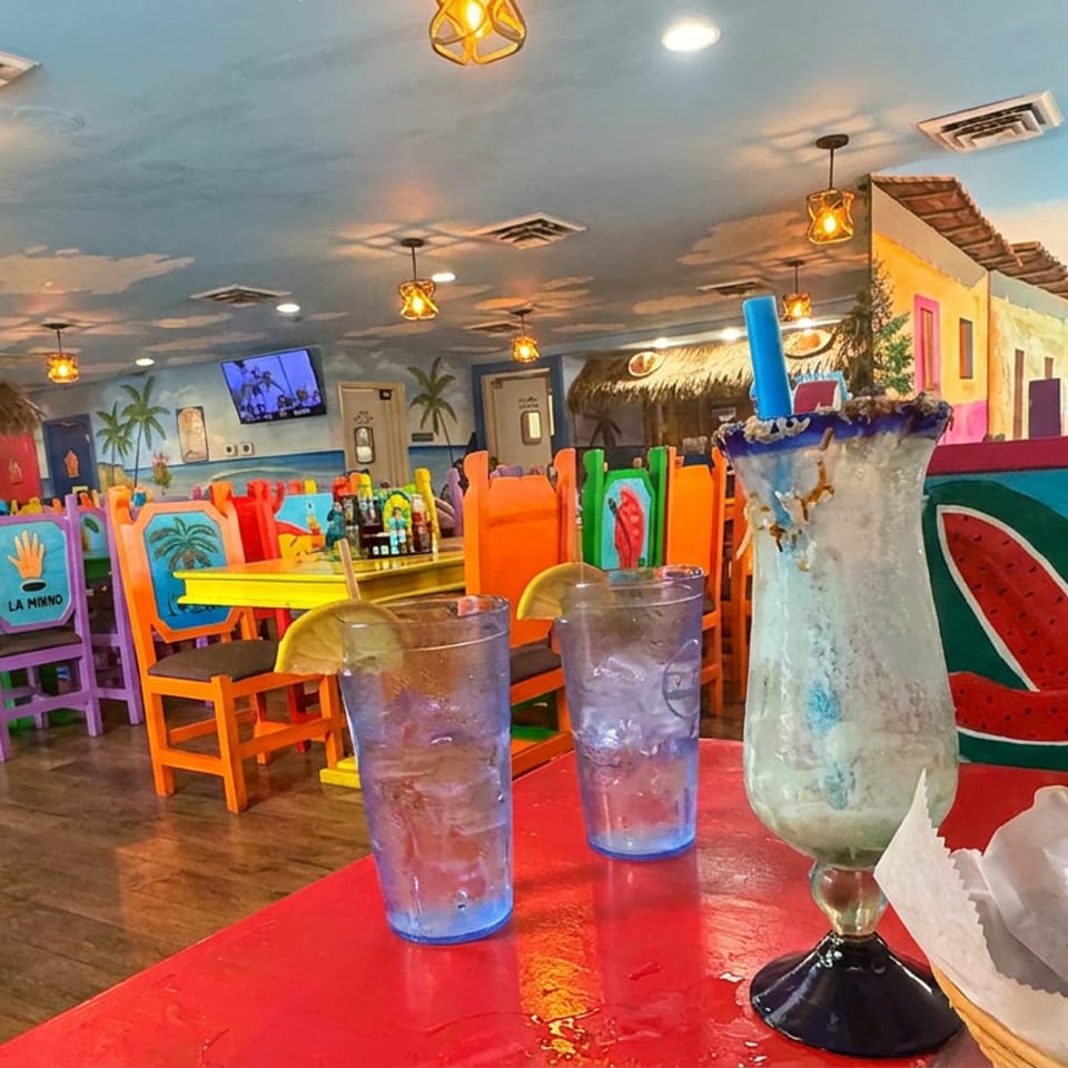 Colorful tropical restaurant interior with mural-painted walls, vivid chairs, two iced drinks, and a large frozen cocktail on a red table.