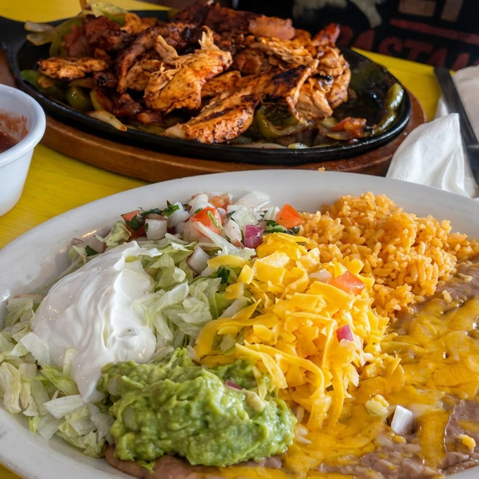 Mexican platter with rice, refried beans, melted cheese, guacamole, sour cream, salad, and grilled shrimp skillet on a yellow table.