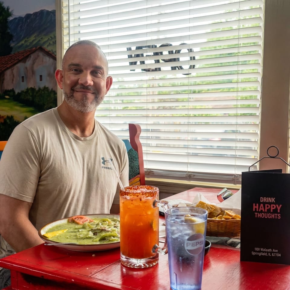 Smiling diner seated at a colorful restaurant table with guacamole, tortilla chips, an orange drink, and iced water.