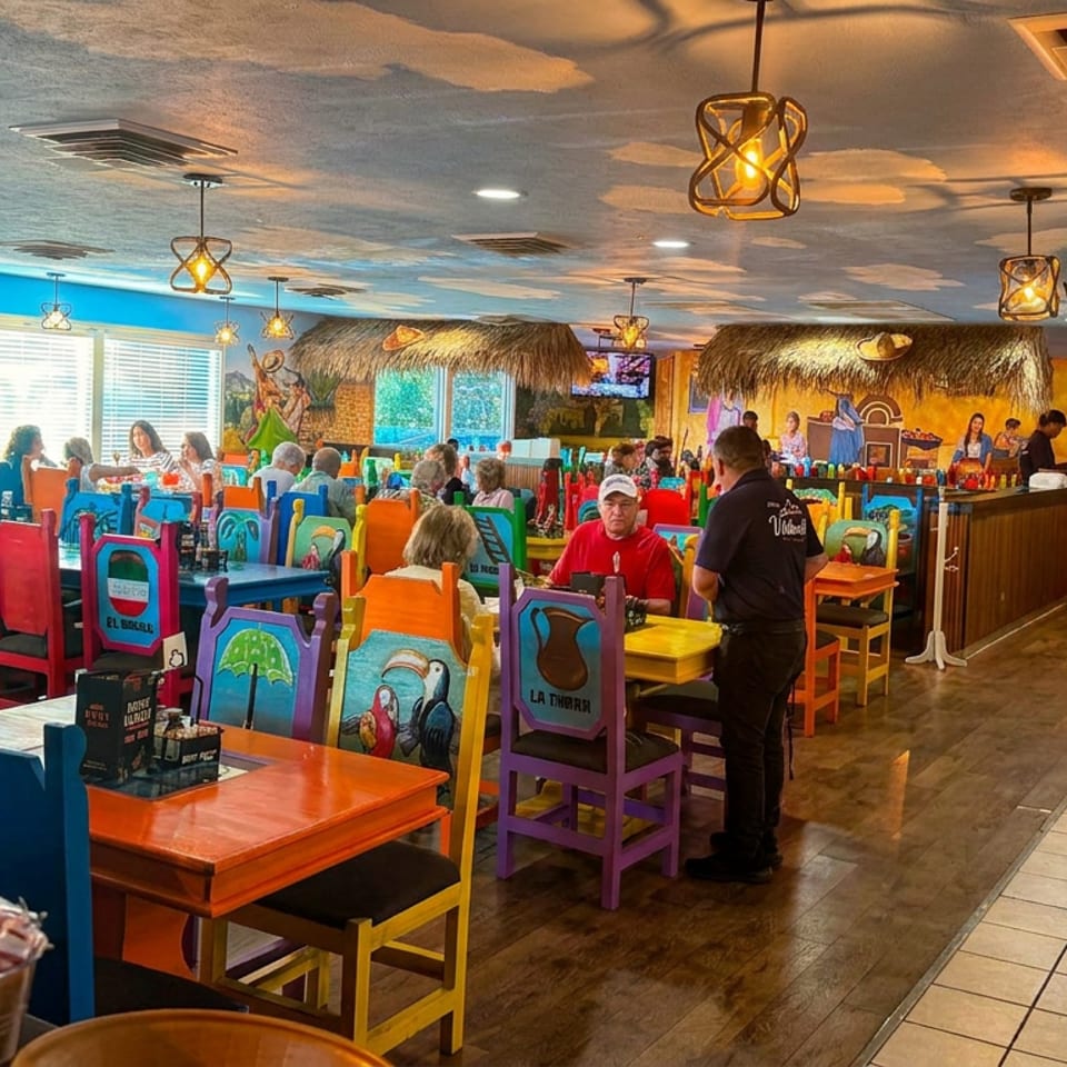 Colorful tropical restaurant dining room with mural-covered walls, thatched bar, pendant lights, and diners seated at vibrant tables.