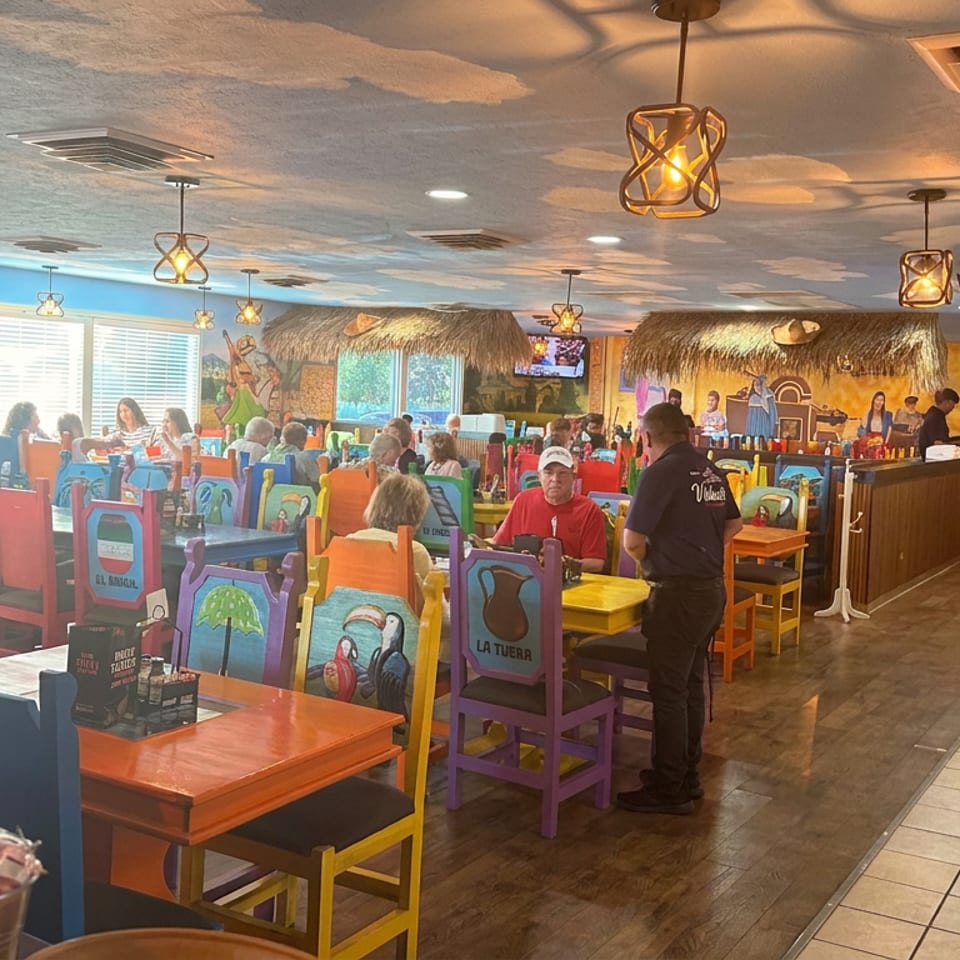Colorful Mexican restaurant dining room with painted chairs, tropical murals, thatched bar, hanging lights, and diners.