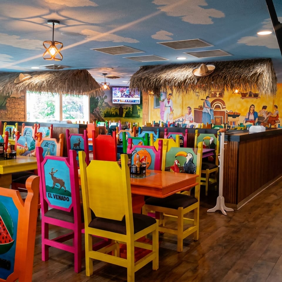 Colorful tropical-themed restaurant dining room with mural walls, thatched roofs, painted chairs, wooden tables, and a service counter.