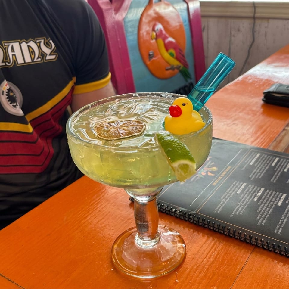Margarita garnished with lime, a dried citrus slice, and rubber duck sits on a restaurant table beside an open menu.