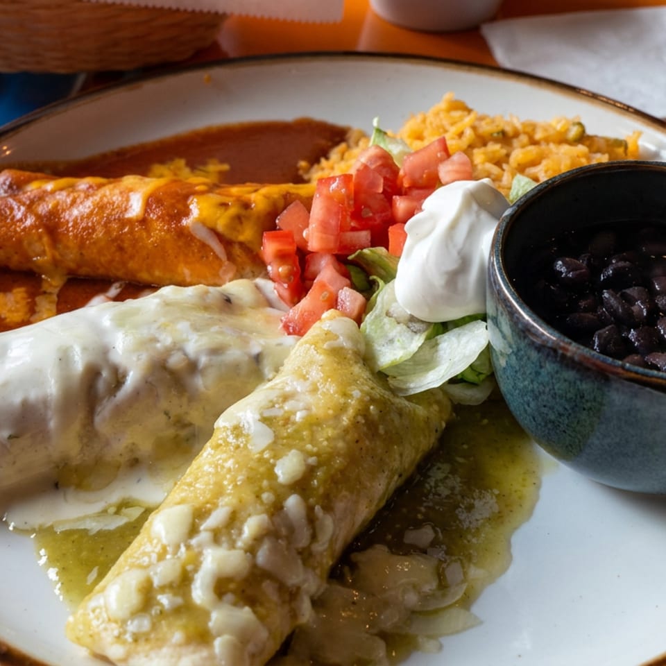Mexican platter with red and green enchiladas, rice, black beans, lettuce, tomatoes, and sour cream on an orange table.